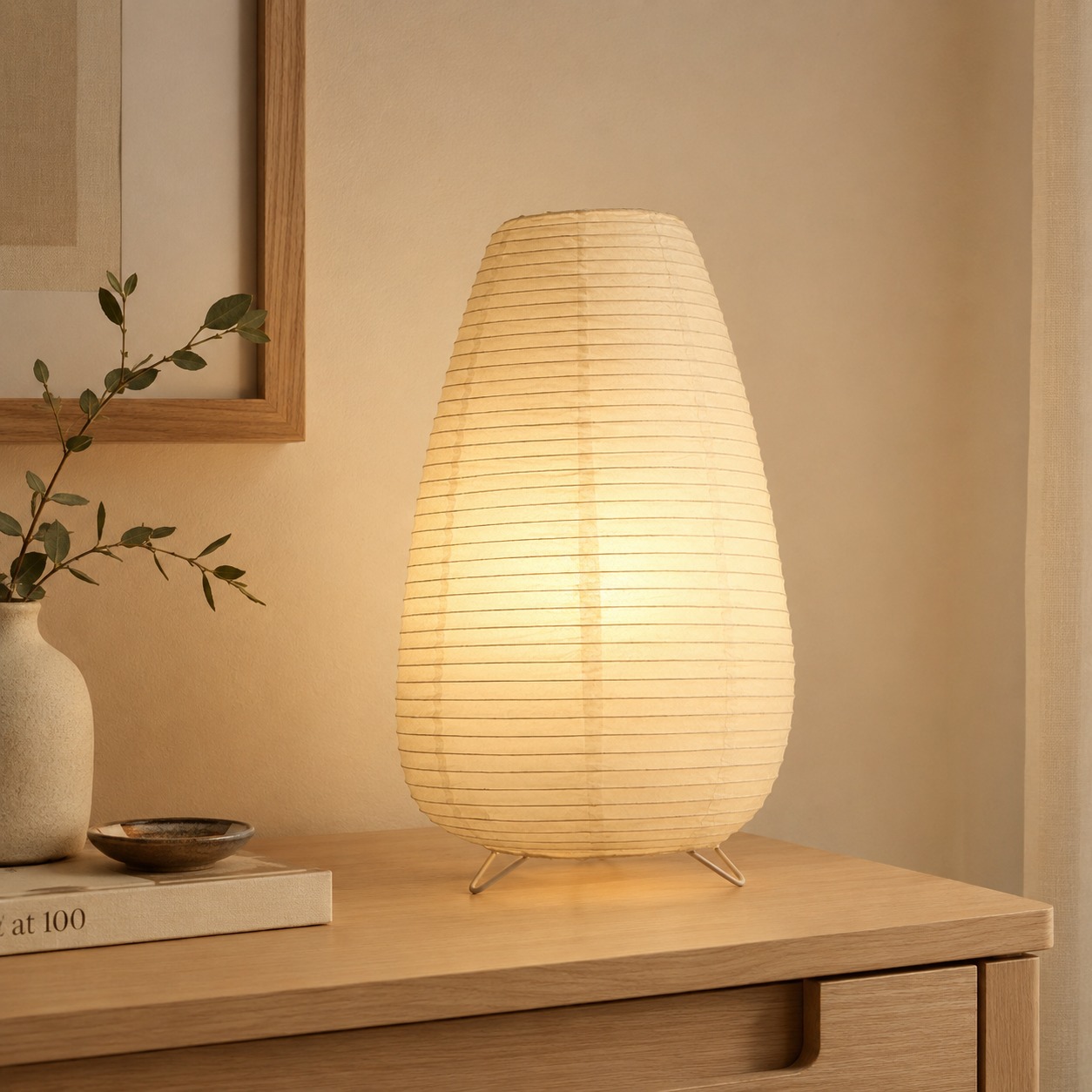 Wooden table lamp on a wooden surface with a soft glow, next to a vase with greenery and a framed picture.